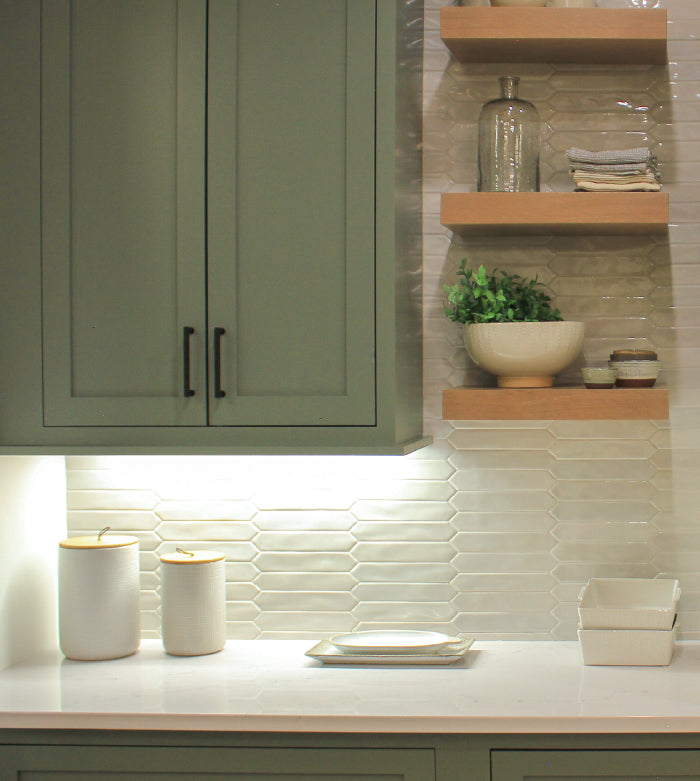 Modern kitchen corner with olive green cabinets, textured white tile backsplash, wooden floating shelves, and minimal countertop decor including ceramic jars and small dishes.