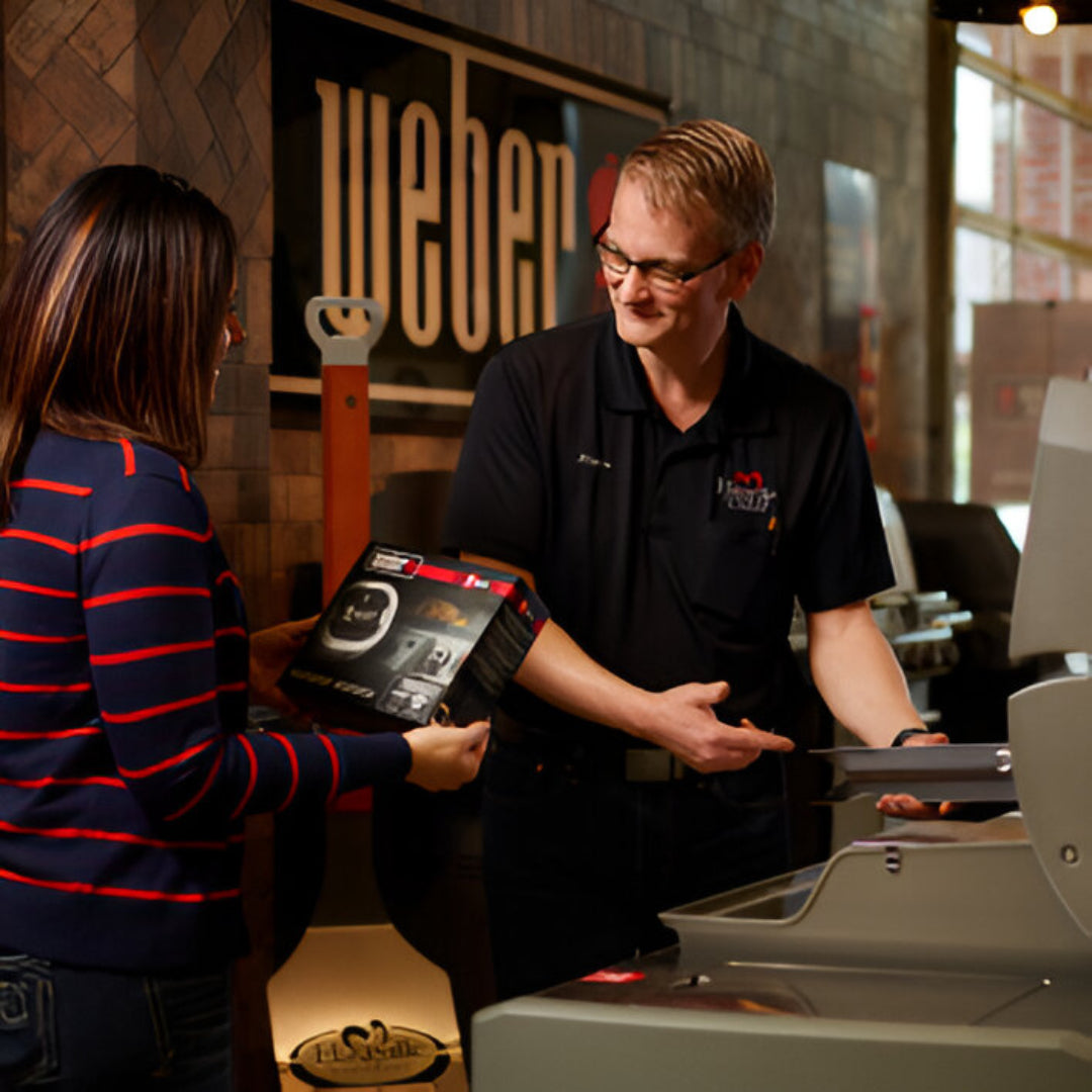 Customer holding a boxed grill accessory while a store employee in a black shirt explains features next to a large stainless steel grill indoors