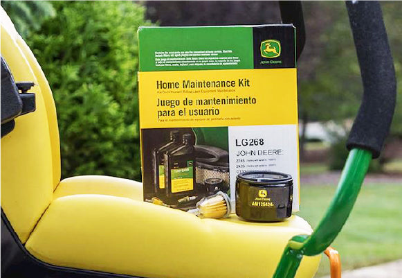 John Deere Home Maintenance Kit in its box with oil filter and fuel filter parts displayed on a yellow tractor seat outdoors with green garden background.