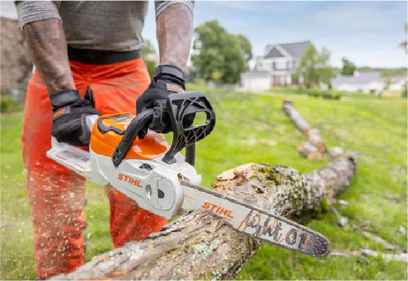 Person wearing gloves and orange pants using a chainsaw to cut a fallen tree branch outdoors in a residential yard.