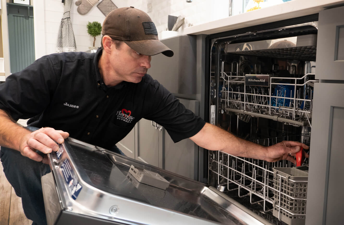 Technician wearing a black shirt and cap inspecting or repairing an open dishwasher door inside a modern kitchen setting.
