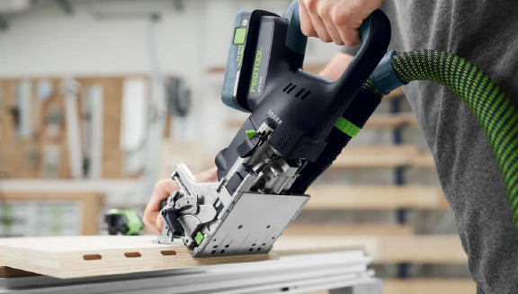 Person using a cordless power tool with a dust extraction hose to cut wooden boards inside a workshop setting.
