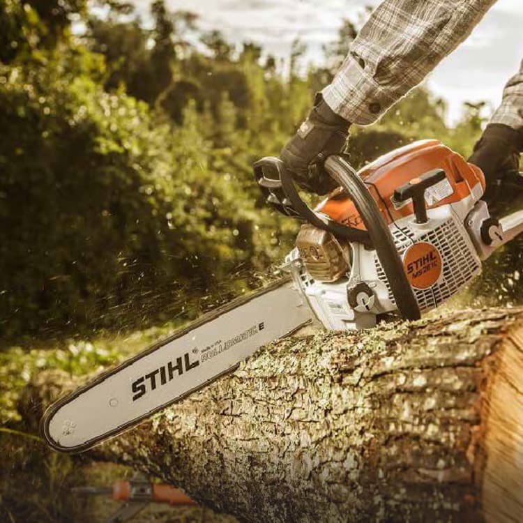 Person wearing gloves and plaid shirt operating a STIHL chainsaw to cut through a large tree trunk outdoors with blurred greenery in the background