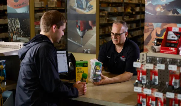Two men engaged in a conversation across a store counter filled with hardware items, with shelves and tools visible in the background.