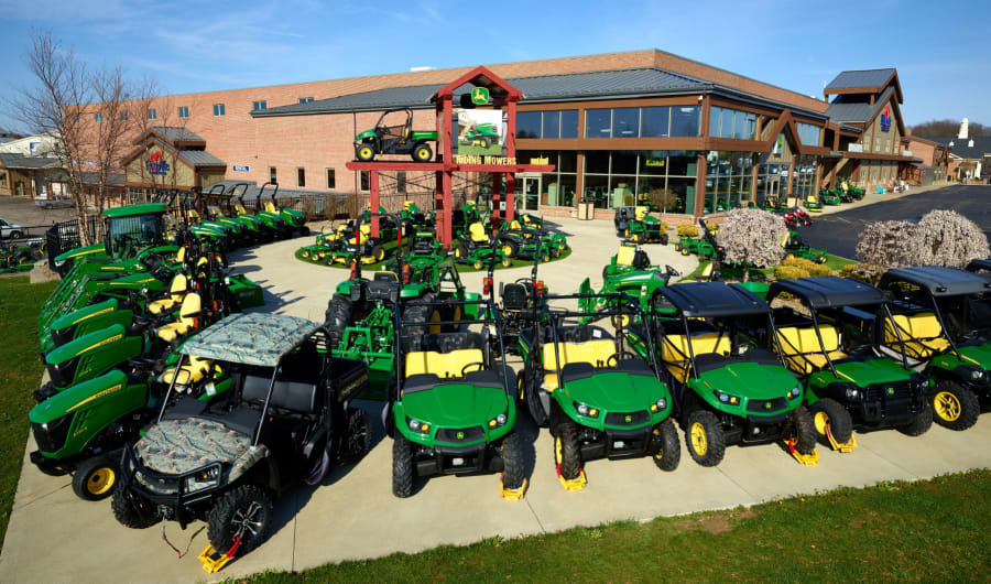 Outdoor display of green and yellow utility vehicles, lawn tractors, and equipment arranged in rows on a concrete patio outside a large brick and glass building.