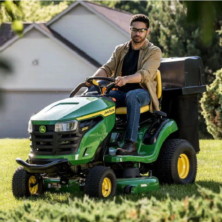Man wearing sunglasses and casual clothing riding a green and yellow lawn tractor on a neatly trimmed lawn in a residential neighborhood