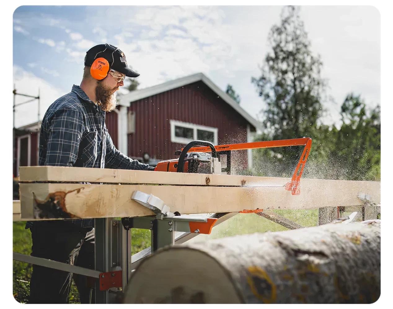 Man wearing protective earmuffs operating an orange sawmill to cut large logs into lumber outdoors near a red building.
