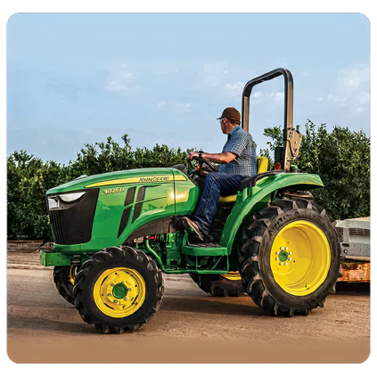 Man wearing jeans and a cap driving a green and yellow John Deere tractor on a dirt path beside dense green bushes under a partly cloudy sky