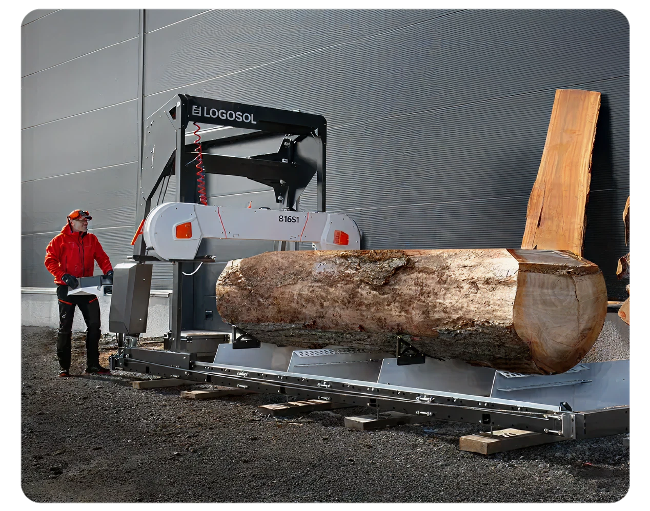 A person wearing protective gear operating a large sawmill machine cutting a thick log outdoors beside a gray building wall.