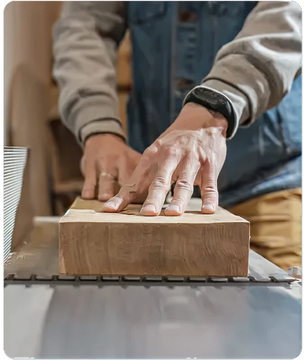 Person wearing a denim vest and gray sleeves guiding a wooden board with both hands on a woodworking table saw in a workshop setting.