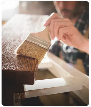 Person applying a coat of varnish on the edge of a wooden surface using a wide paintbrush in a workshop setting