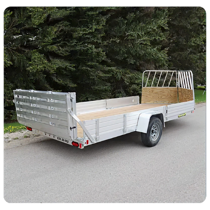 Open aluminum flatbed trailer with metal rails and wooden floor panels parked on a paved road near dense green trees.