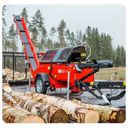 Red heavy-duty wood processor machine with conveyor belt and trailer hitch, positioned outdoors near stacked logs and a forested area.