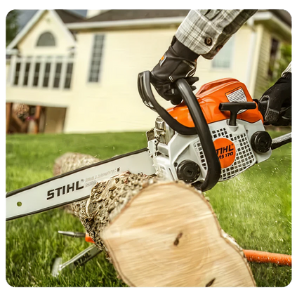 Person wearing protective gloves operating an orange and white chainsaw to cut a log of wood outdoors with a house in the background