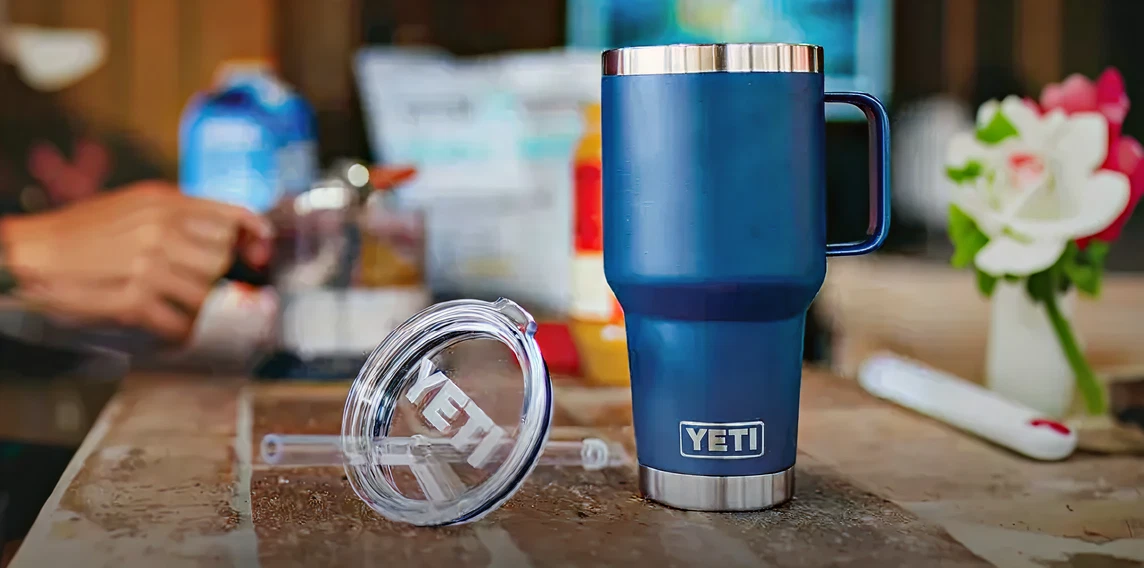 Blue insulated YETI tumbler with a handle and clear lid resting on a wooden table in a home setting with blurred background items and a white flower in a vase.