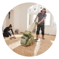 Man sanding a hardwood floor with a large floor sander while a woman works on the floor edges in a bright room with windows.