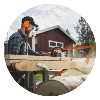 Man wearing protective earmuffs operating a chainsaw to cut a large wooden log outdoors near a red barn during daytime.